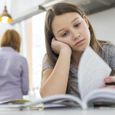 Girl studying at table with mother standing in background