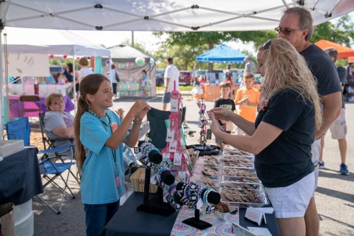 Girl behind table in tent selling bracelets and cookies with man and woman customers at Acton Children's Business Fair